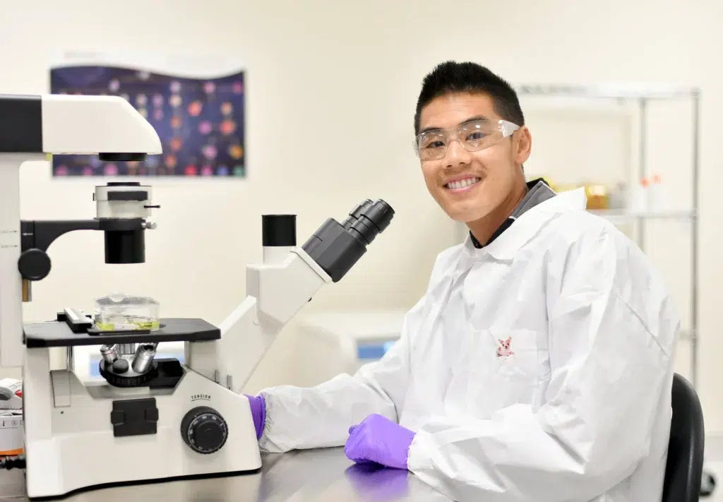 Young researcher in a lab coat and safety glasses beside a microscope, healthcare and life sciences photography by Josh Edelson