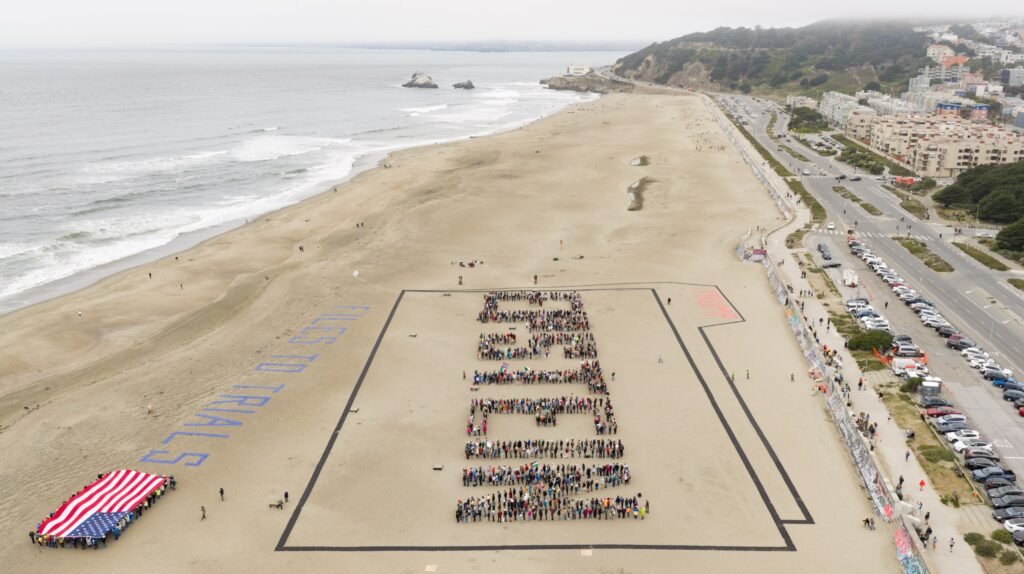 Aerial drone view over the Pacific Ocean of a human-banner protest at Ocean Beach, San Francisco, spelling EPSTEIN FILES TO TRIALS in the sand