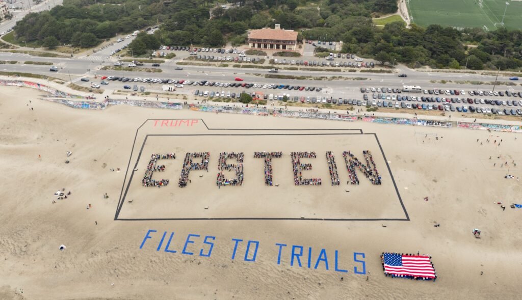 Aerial drone photo of the EPSTEIN FILES TO TRIALS human banner and a giant American flag on Ocean Beach, San Francisco, with the Great Highway and Beach Chalet behind