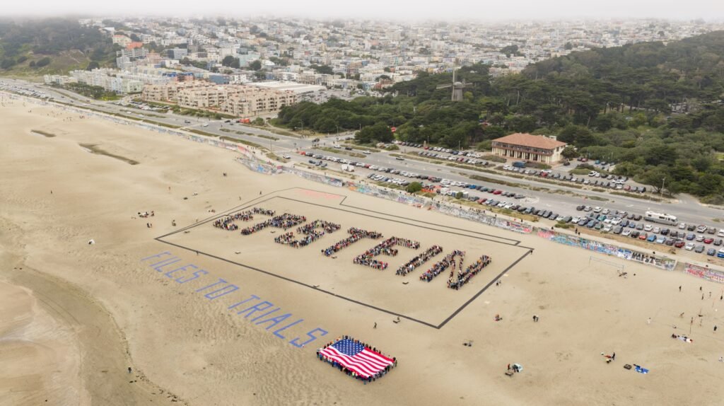 Aerial drone photo of more than 1,000 people forming a human banner that spells EPSTEIN on Ocean Beach in San Francisco, with the Sunset District and fog behind