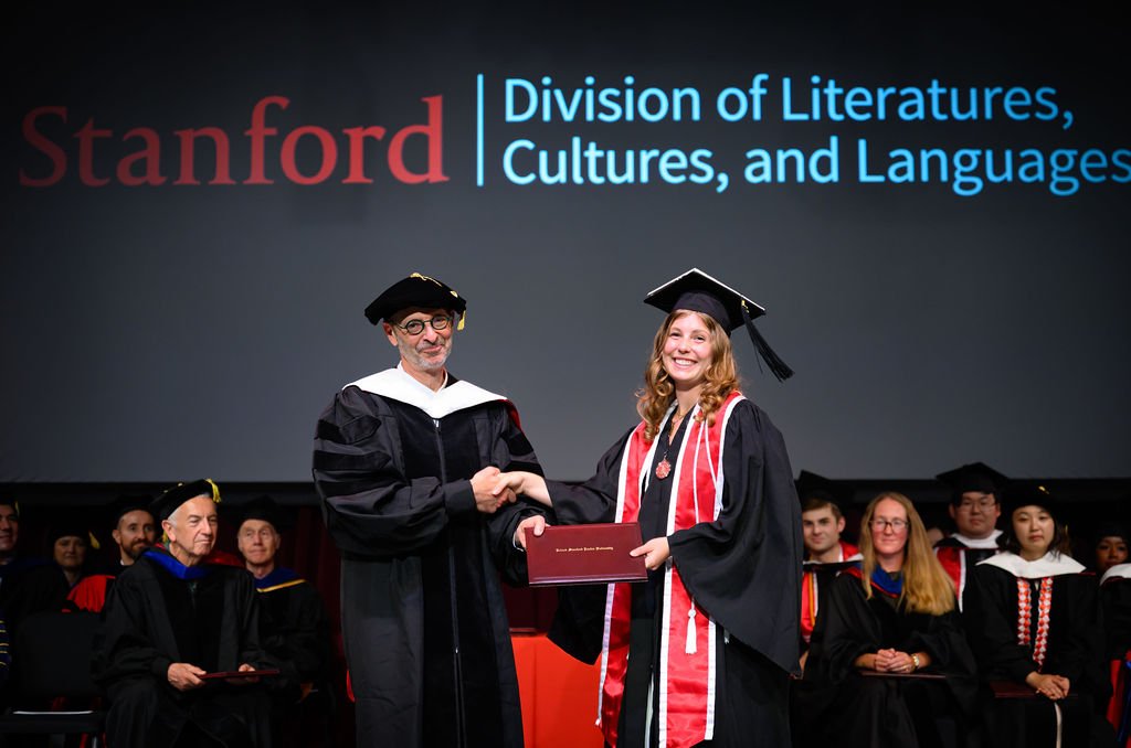 A graduate shakes hands and receives a diploma on stage under the Stanford DLCL backdrop at commencement