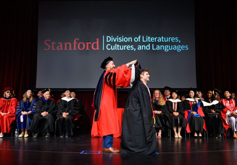 A faculty member hoods a doctoral graduate on stage beneath the Stanford Division of Literatures, Cultures, and Languages backdrop at the DLCL commencement