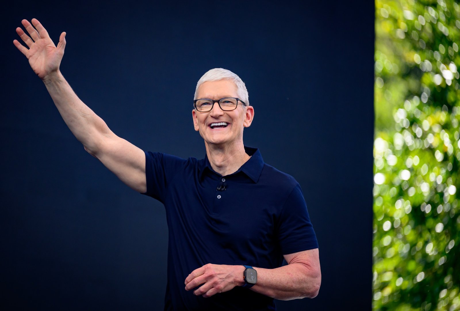 Apple CEO Tim Cook smiling and waving on stage at the WWDC 2026 keynote, with green Apple Park foliage behind him