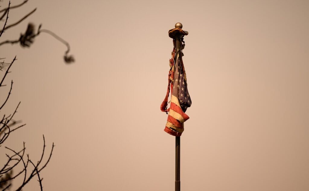 A burned American flag rests atop a flagpole at a burned home during the Old Trails Fire in Spokane, Washington, August 4, 2026. (Josh Edelson / AFP)