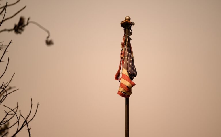 A burned American flag rests atop a flagpole at a burned home during the Old Trails Fire in Spokane, Washington, August 4, 2026. (Josh Edelson / AFP)